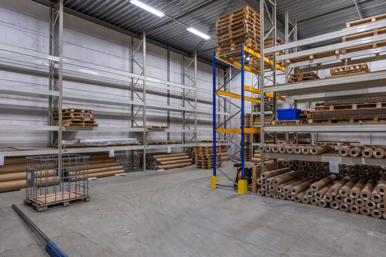 Interieur van magazijn met metalen planken met houten pallets en rollen bruin papier; sommige planken zijn gedeeltelijk leeg.