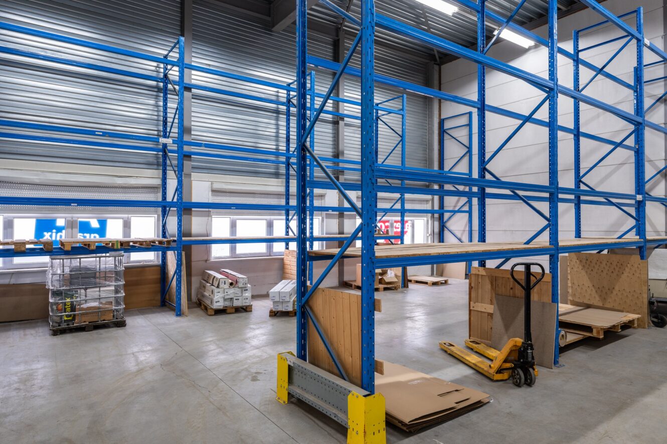 Lege blauwe metalen magazijnplanken met enkele pallets, dozen en een gele pallethefwagen op een betonnen vloer in een goed verlichte industriële opslagruimte.