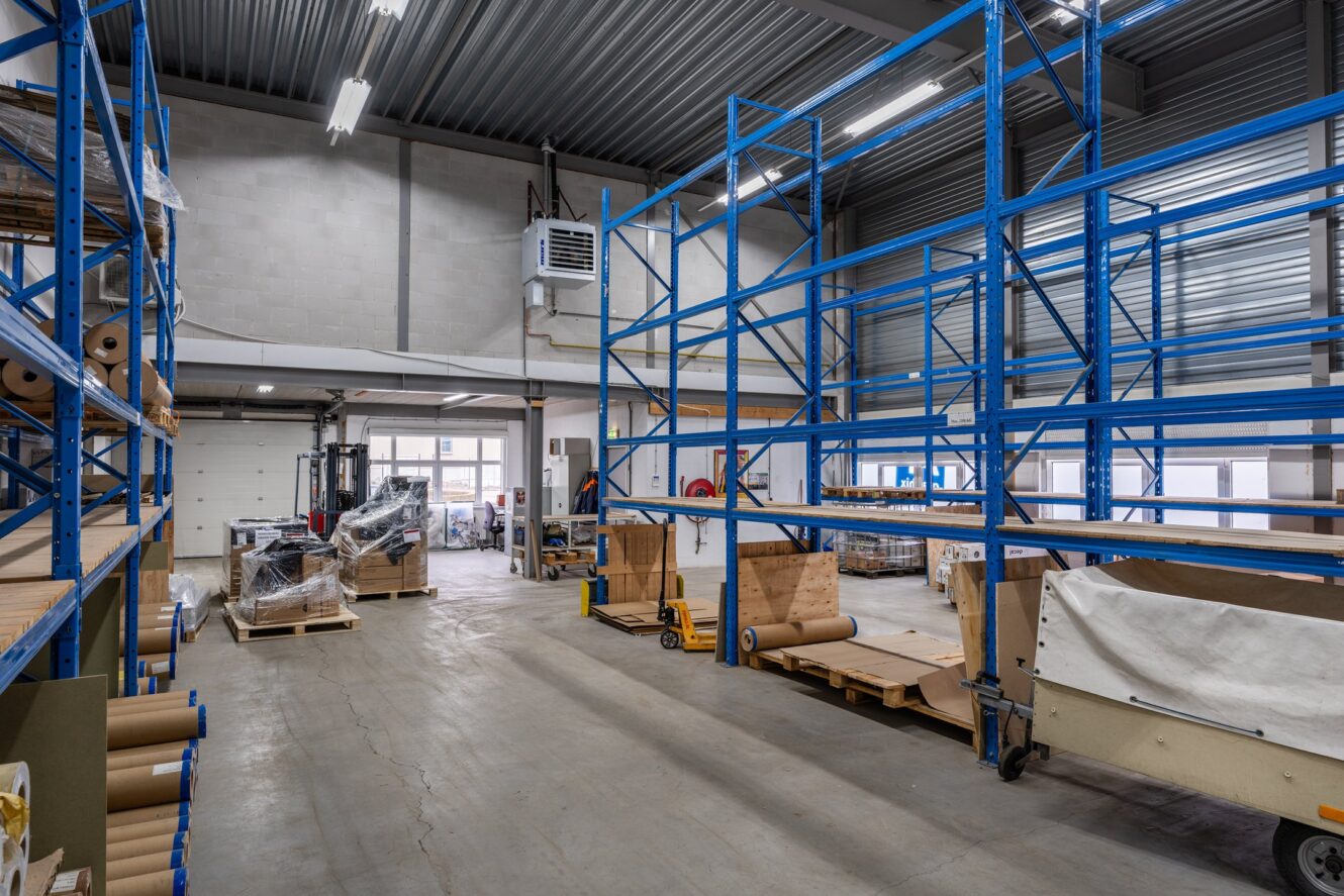 Industrieel magazijninterieur met lege blauwe metalen opslagrekken, verspreide pallets en verpakkingsmateriaal op de vloer onder heldere overheadverlichting.