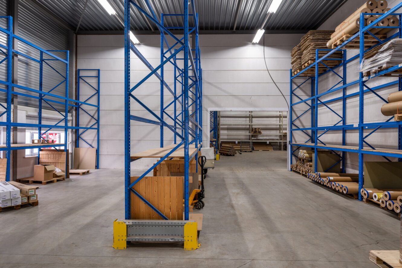 Groot magazijninterieur met blauwe metalen rekken, opgerolde materialen, kartonnen dozen en enkele houten pallets op de betonnen vloer onder heldere plafondverlichting.