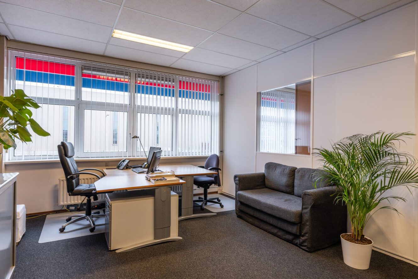 Een modern kantoor met twee bureaus en stoelen, een zwarte bank, planten in potten, grote ramen met luxaflex en witte muren.