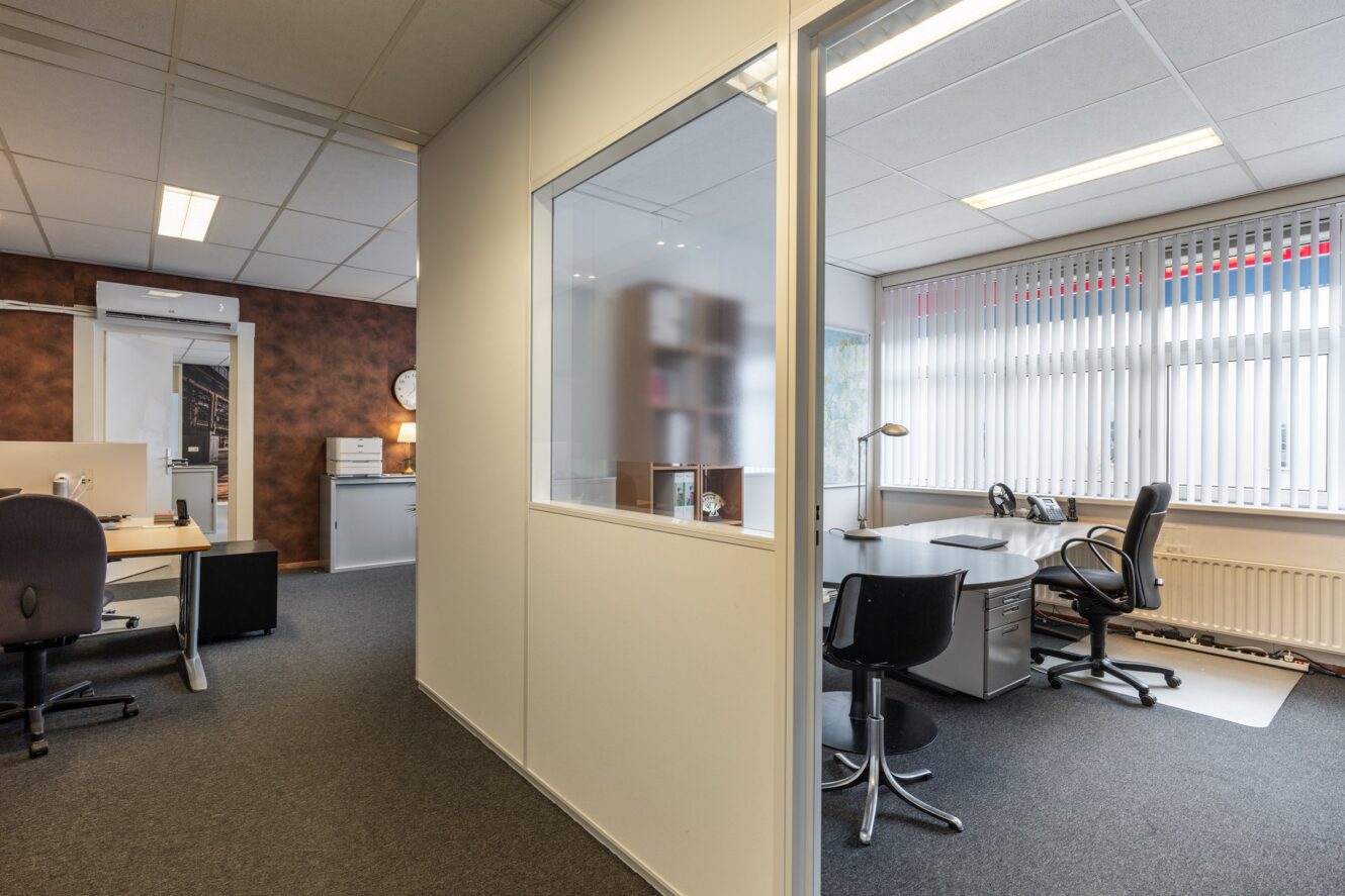 Moderne kantoorruimte met ingedeelde kamers, bureaus, stoelen, computers en grote ramen met verticale jaloezieën die natuurlijk licht binnenlaten.