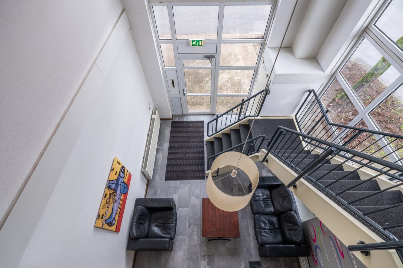 Zicht van bovenaf op de lobby van een klein gebouw met zwartleren banken, een houten tafel, een trap en een grote toegangsdeur met een raam en een bordje met de uitgang erboven.