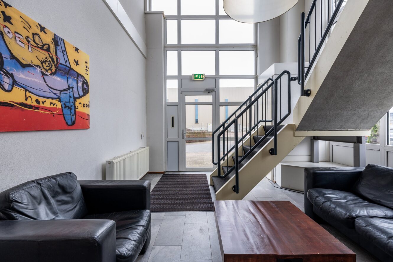 Een moderne lobby met zwartleren banken, een houten salontafel, abstracte muurkunst en een trap naar boven naast grote ramen en een glazen deur.