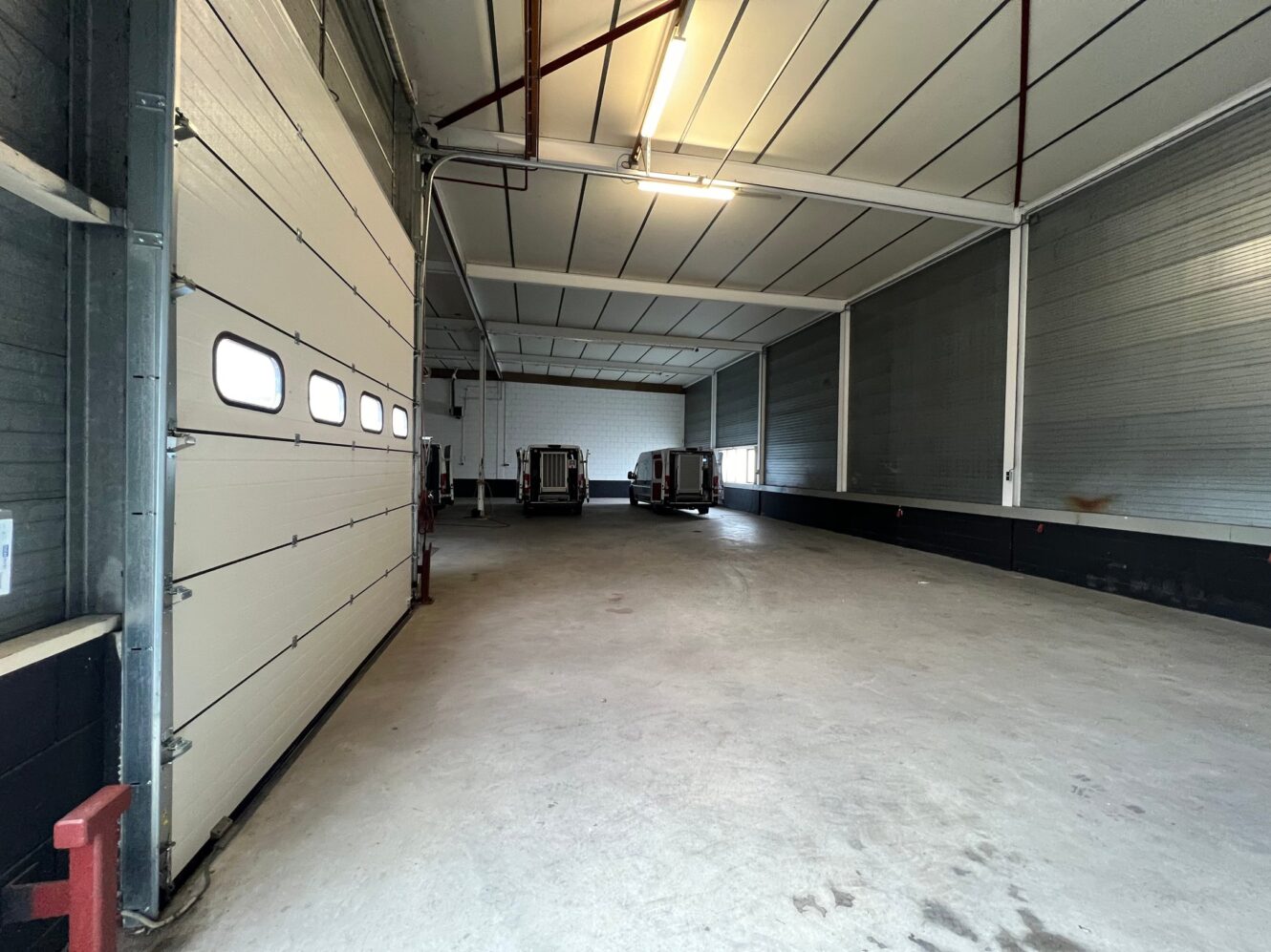 Interieur van een leeg magazijn met betonnen vloer, metalen muren, hoog plafond en twee laadkades met achteraan trailers zichtbaar aan het einde.