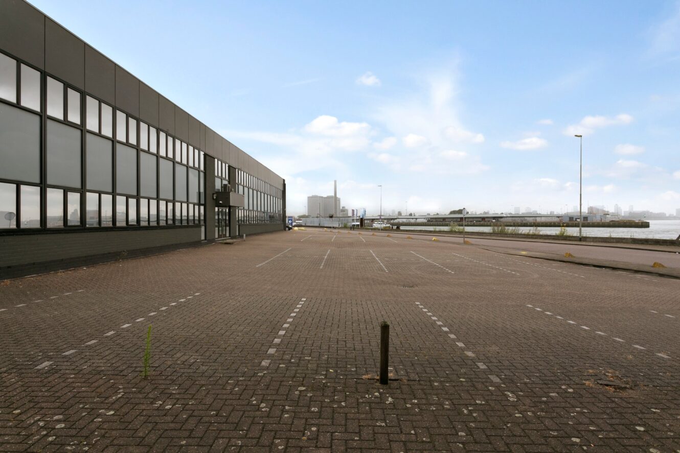 Lege parkeerplaats naast een groot industrieel gebouw met water en fabrieken in de verte zichtbaar onder een blauwe lucht met verspreide wolken.