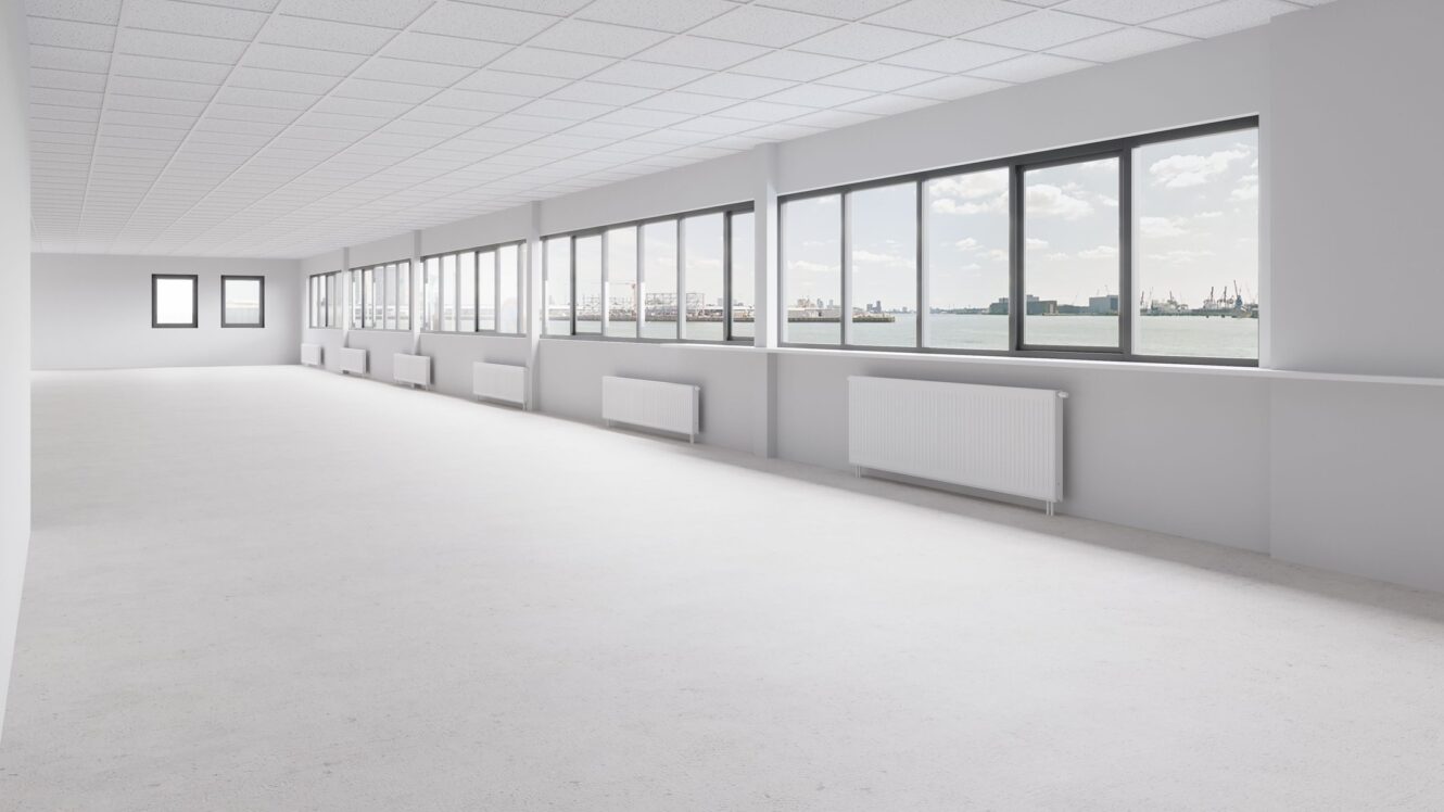 Grote, lege, witte kamer met veel ramen, radiatoren onder elk raam en uitzicht op water en een industriële haven buiten.