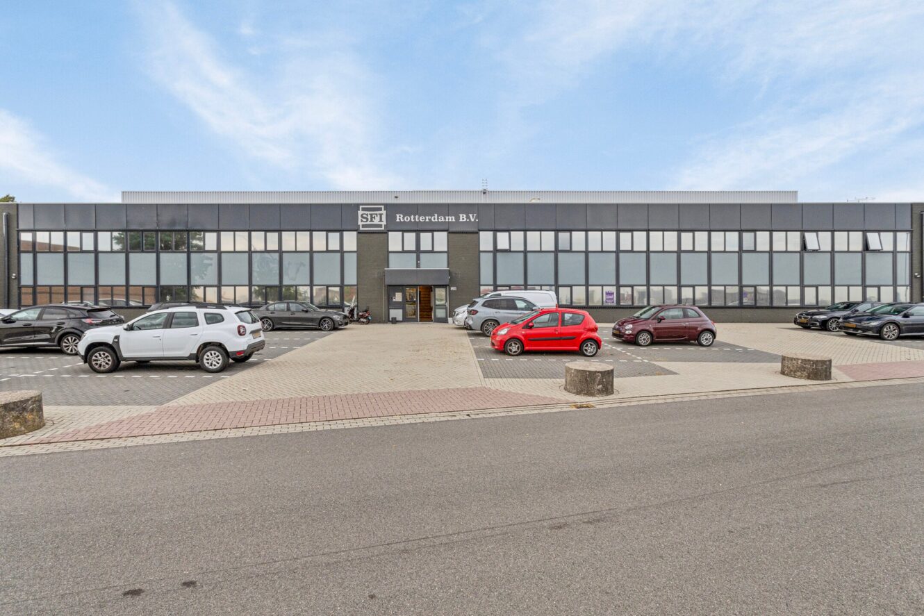Een bedrijfsgebouw met de naam SPT Rotterdam B.V. met een aantal geparkeerde auto's ervoor op een verhard parkeerterrein, onder een gedeeltelijk bewolkte hemel.