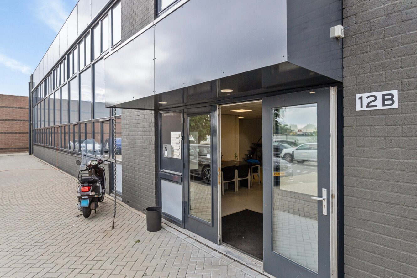 Ingang van een modern commercieel gebouw met glazen ramen, open dubbele deuren, een scooter buiten geparkeerd en een bord met 12B aan de muur.