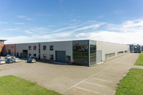 Groot, modern industrieel pakhuis met glazen ramen, verschillende geparkeerde auto's en een grotendeels lege parkeerplaats onder een strakblauwe hemel.
