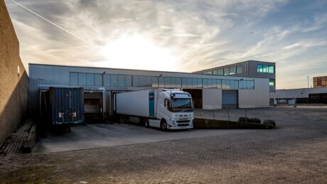 Een witte vrachtwagen staat geparkeerd bij een laadperron buiten een modern industrieel pakhuis onder een gedeeltelijk bewolkte hemel bij zonsondergang.