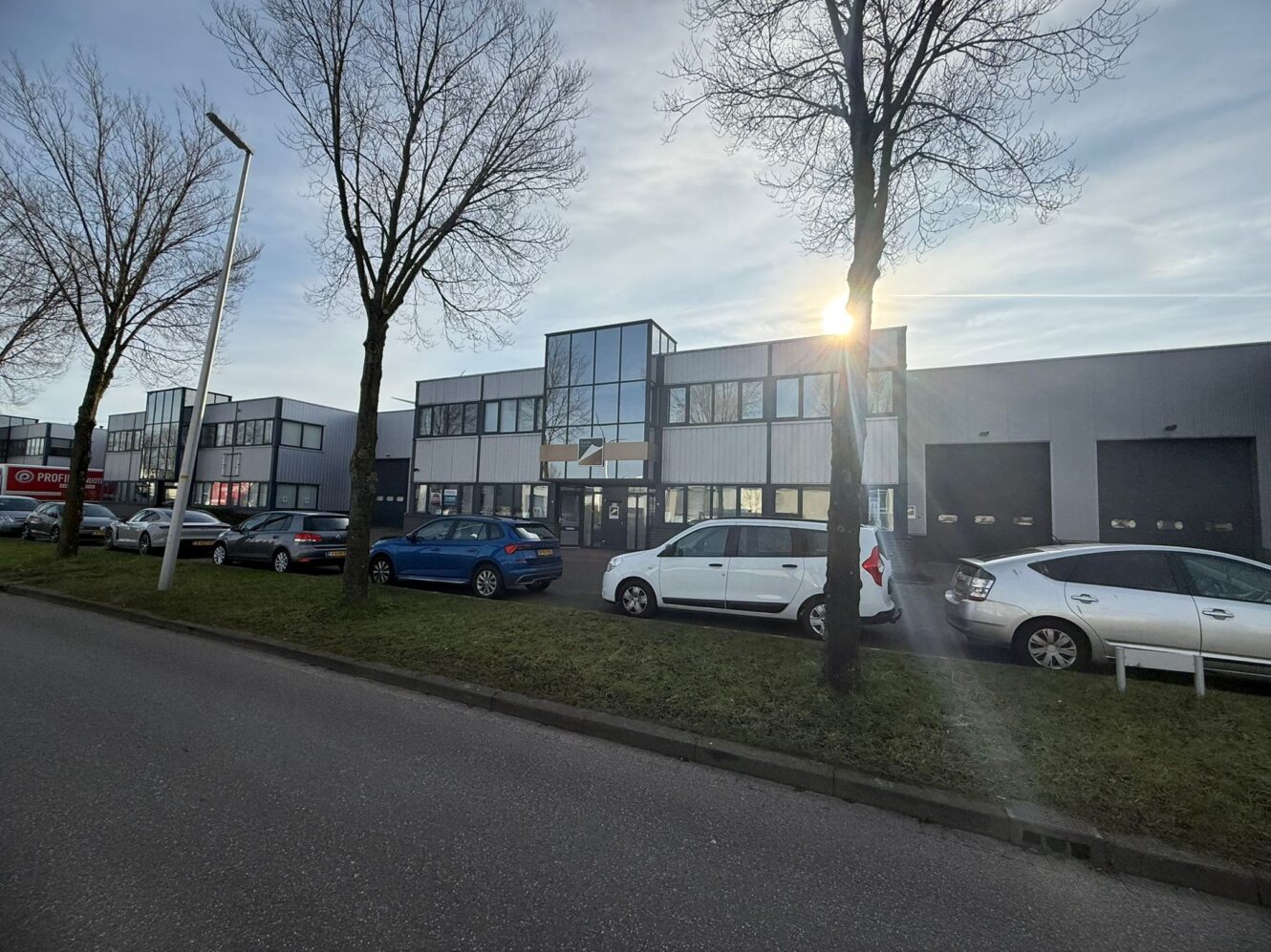 Een rij geparkeerde auto's omzoomt de straat voor een modern, grijs commercieel gebouw met grote ramen en bladerloze bomen.