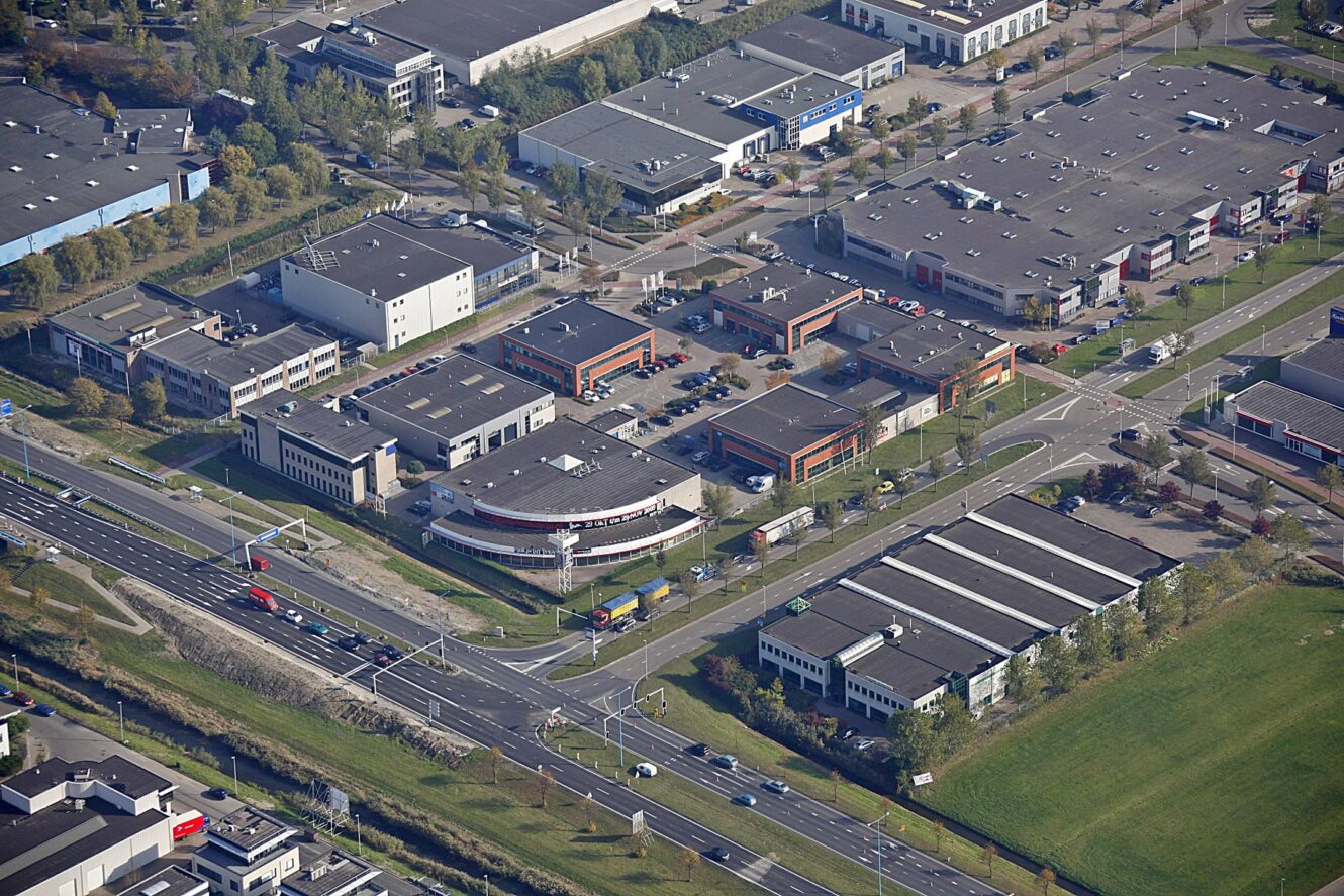 Luchtfoto van een industrieel gebied met meerdere magazijnen, kantoorgebouwen, parkeerplaatsen en wegen met auto's en vrachtwagens. Grasvelden en bomen omzomen het complex.