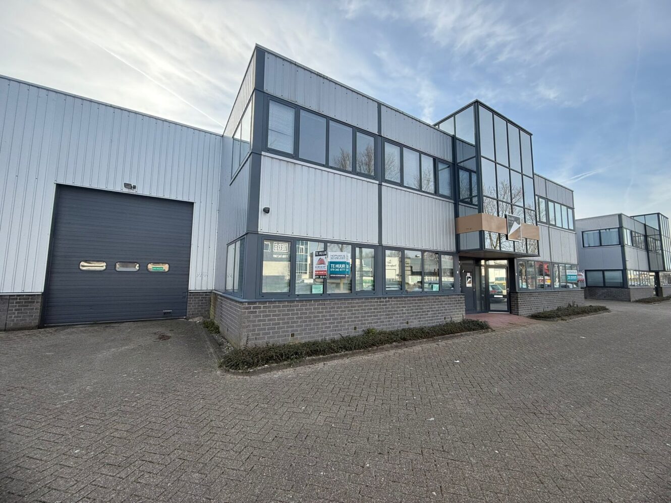Modern industrieel gebouw met grote ramen, een glazen ingang, zilveren lambrisering en een aangrenzende laadperrondeur, gelegen op een verhard terrein onder een blauwe hemel.
