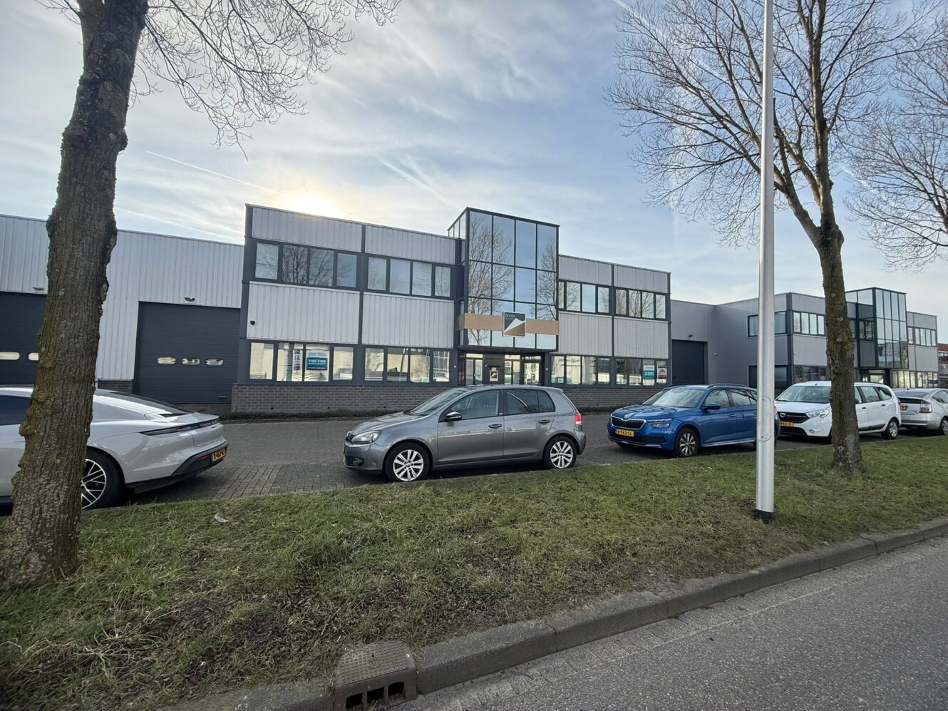 Een rij auto's geparkeerd voor een modern industrieel gebouw met grote ramen en een grijze gevel op een bewolkte dag.