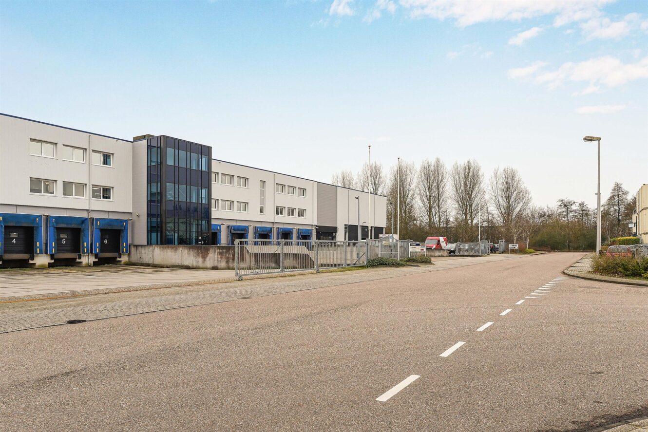 Een modern industrieel gebouw met laadkades en een omheind terrein, grenzend aan een lege straat en omringd door bomen onder een gedeeltelijk bewolkte hemel.