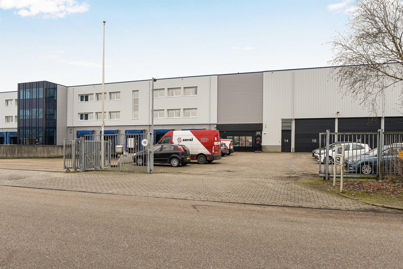 Een modern industrieel gebouw met grote ramen, meerdere garagedeuren en een parkeerplaats met meerdere auto's en een rood busje, gezien vanaf de straat.