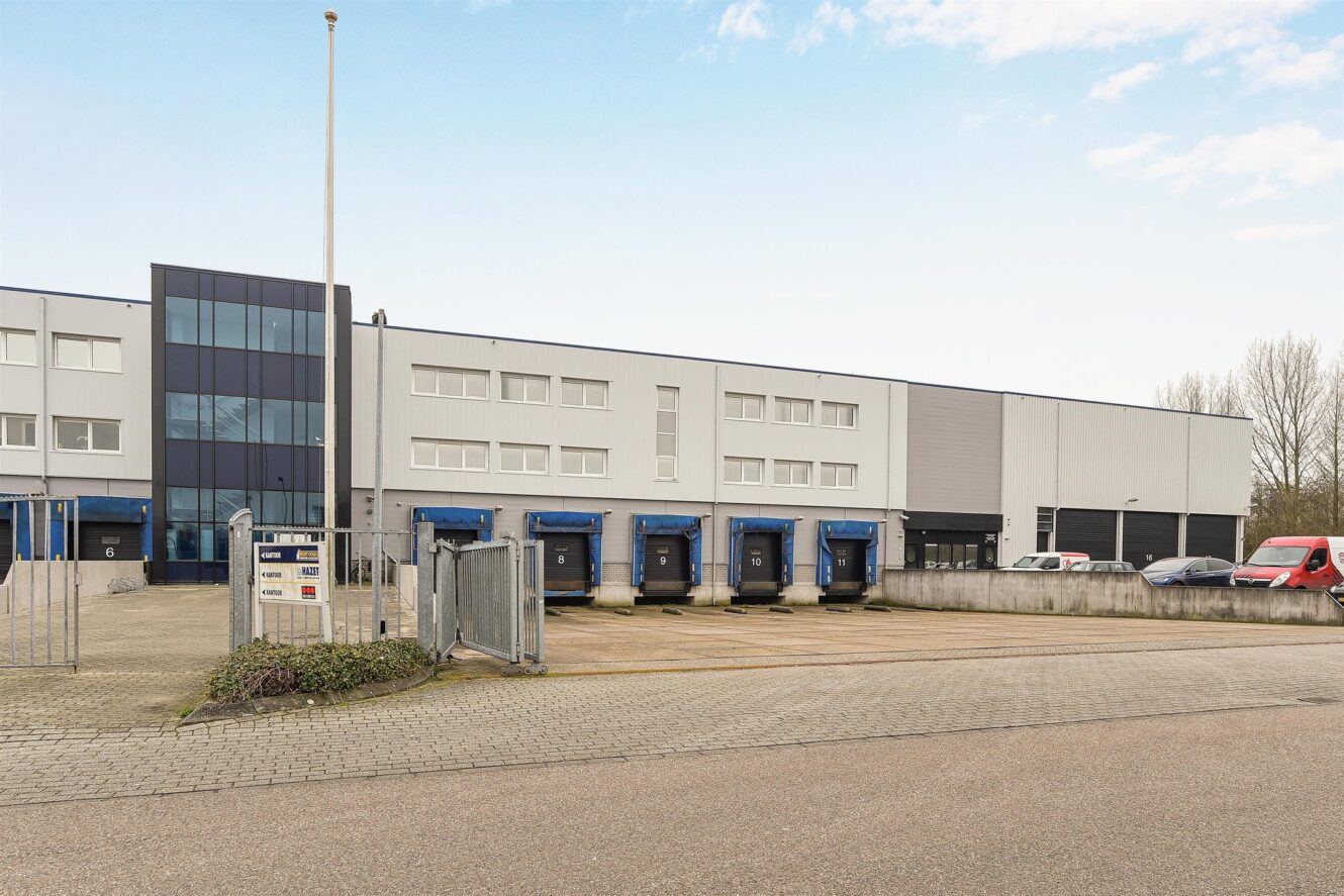 Modern industrieel pakhuis met meerdere laaddocks, glazen ingang en verschillende geparkeerde voertuigen op een verhard terrein onder een gedeeltelijk bewolkte hemel.