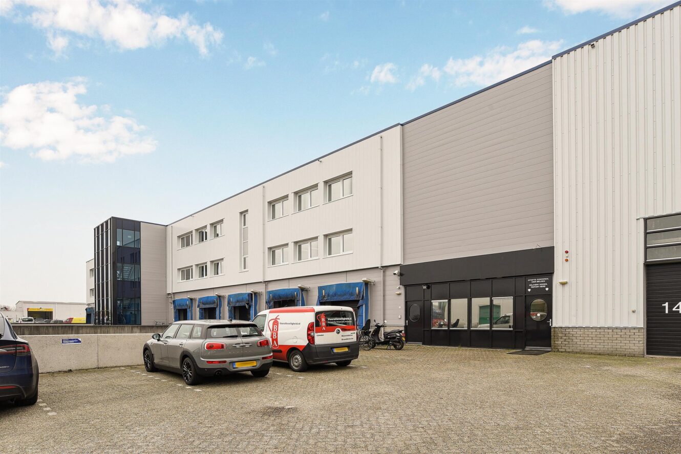 Een modern industrieel gebouw met een aantal geparkeerde auto's ervoor, met grote ramen, laadkades en een kleine glazen ingang.