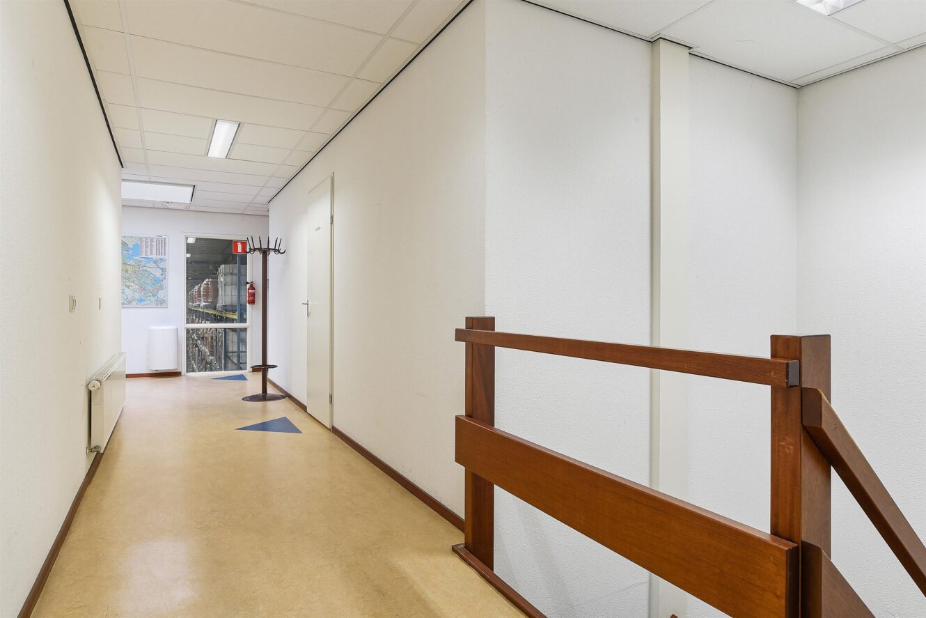 Een hal met beige vloeren, witte muren, een houten balustrade aan de rechterkant en een kapstok en brandblusser bij een glazen deur aan het einde.