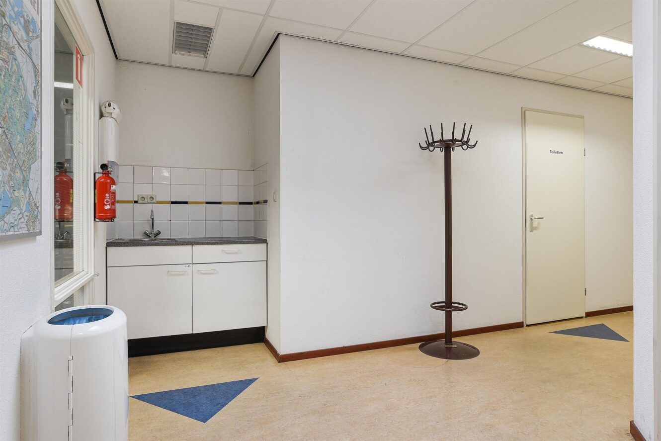 Een klein keukentje met een gootsteen en kastjes, een kapstok, een gesloten deur met het label Toiletten, een brandblusser en een prullenbak in een schone gang.