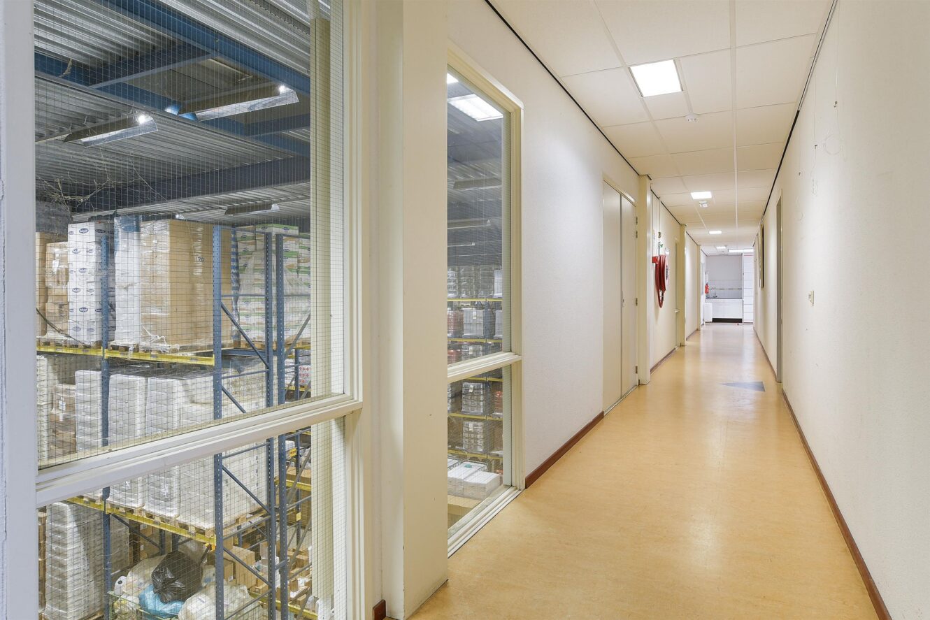 Een hal met beige vloeren en witte muren, met ramen die uitkijken op een magazijn met rekken en gestapelde dozen; fluorescerende lampen aan het plafond.