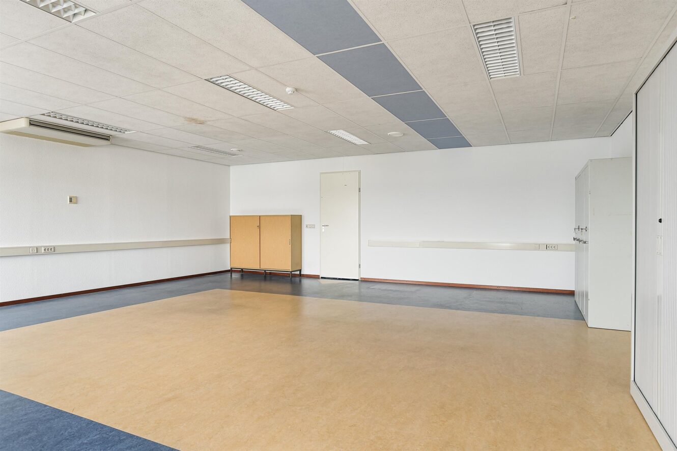 Lege kantoorkamer met een beige en blauwe vloer, witte muren, plafondverlichting, een gesloten deur, een kast en een opbergmeubel tegen de muren.