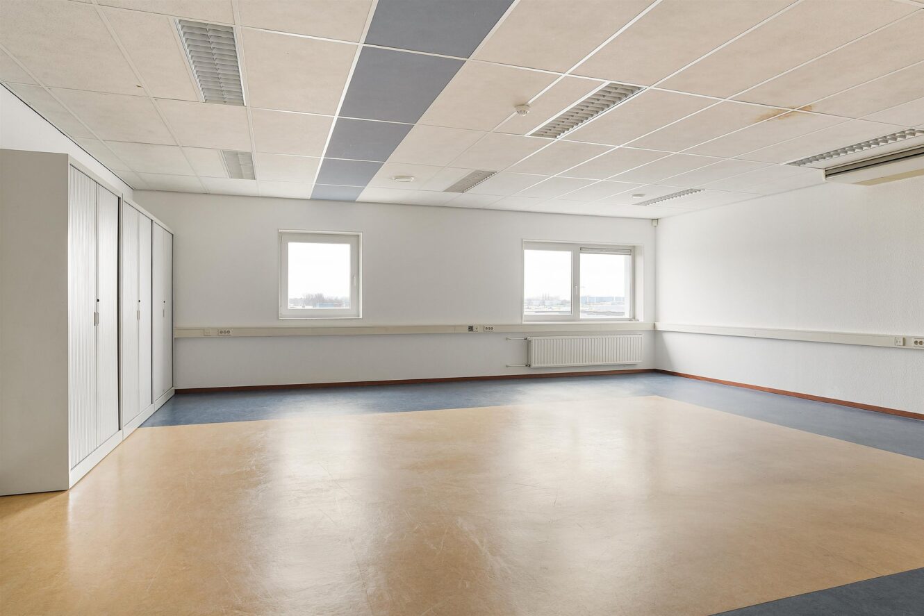 Lege kamer met lichtgekleurde muren, blauwe en beige vloer, twee ramen en tl-verlichting aan het plafond. Ingebouwde kasten staan aan de linkerkant van de kamer.