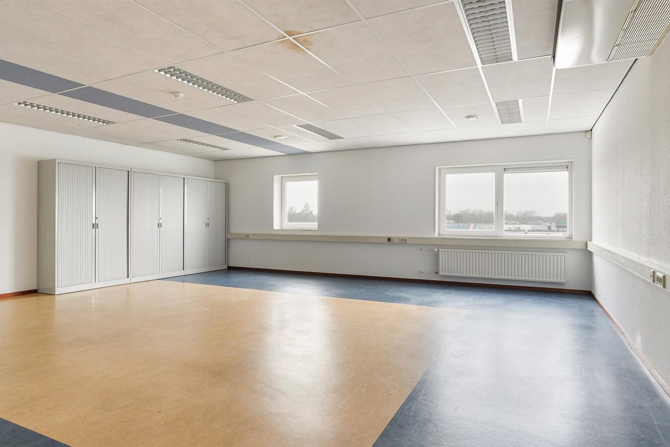 Lege kamer met beige en blauwe vloeren, witte plafondtegels, tl-verlichting, twee ramen en een rij witte opbergkasten tegen de achterwand.