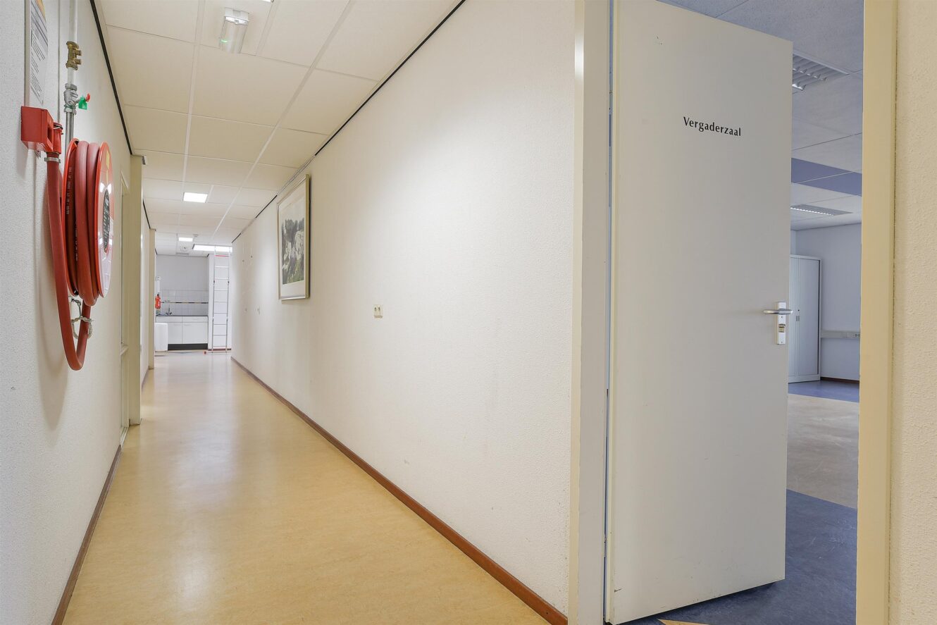 Een hal met beige vloeren, witte muren, een brandslanghaspel aan de linkerkant en een open deur met het label Vergaderzaal aan de rechterkant.