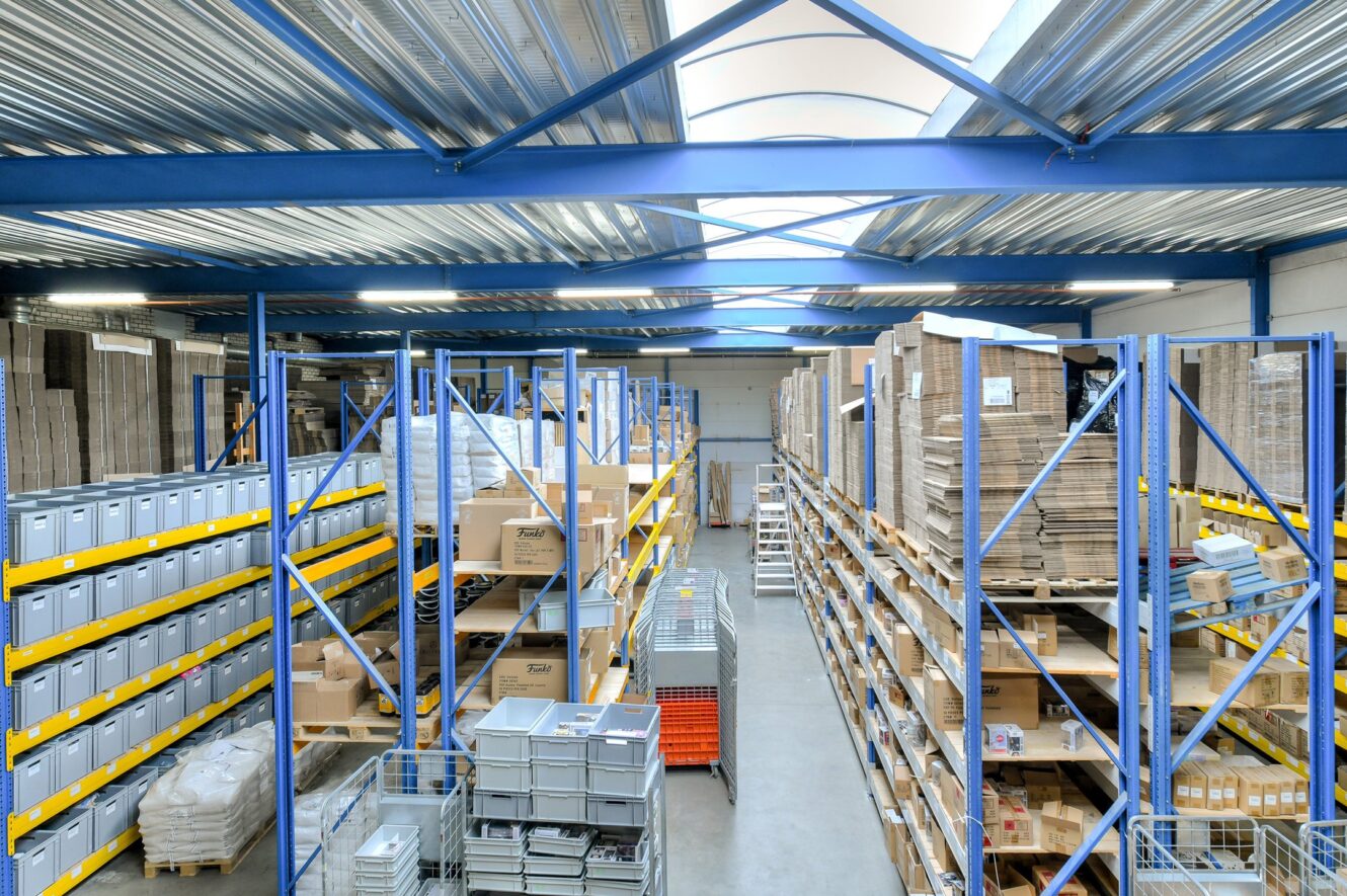 Goed verlicht magazijninterieur met hoge metalen rekken vol dozen en verschillende artikelen, georganiseerd in duidelijk gelabelde secties langs een centraal gangpad.