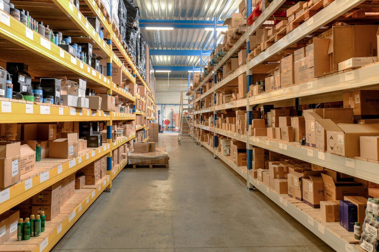Een magazijngang met hoge rekken vol kartonnen dozen en verschillende producten aan beide kanten, onder tl-verlichting.