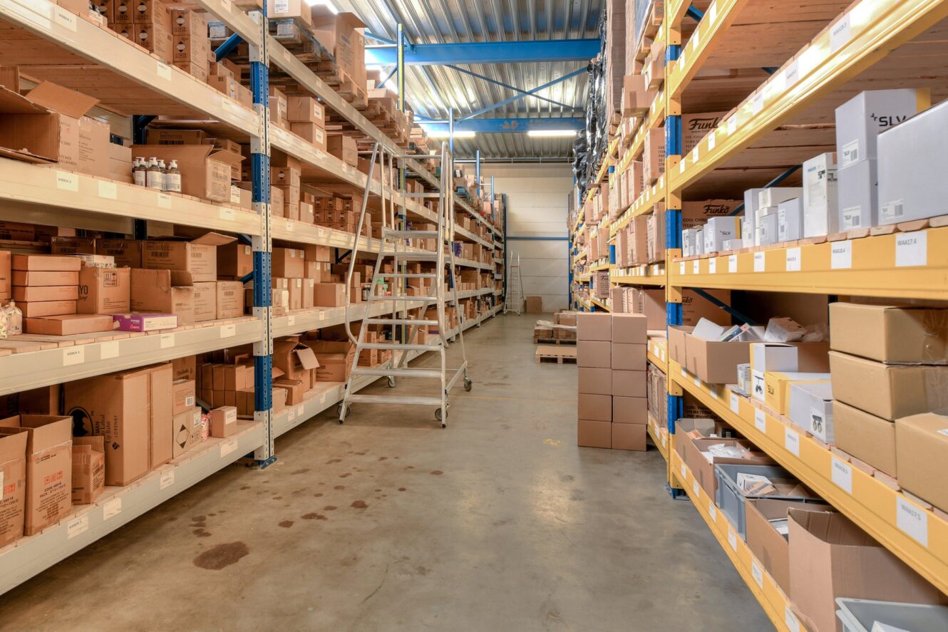 Een magazijngang met rekken vol verschillende kartonnen dozen en een metalen ladder in het midden.