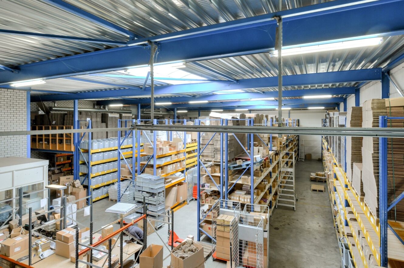 Groot magazijn met metalen rekken gevuld met dozen en pakjes, betonnen vloer en bovenverlichting.