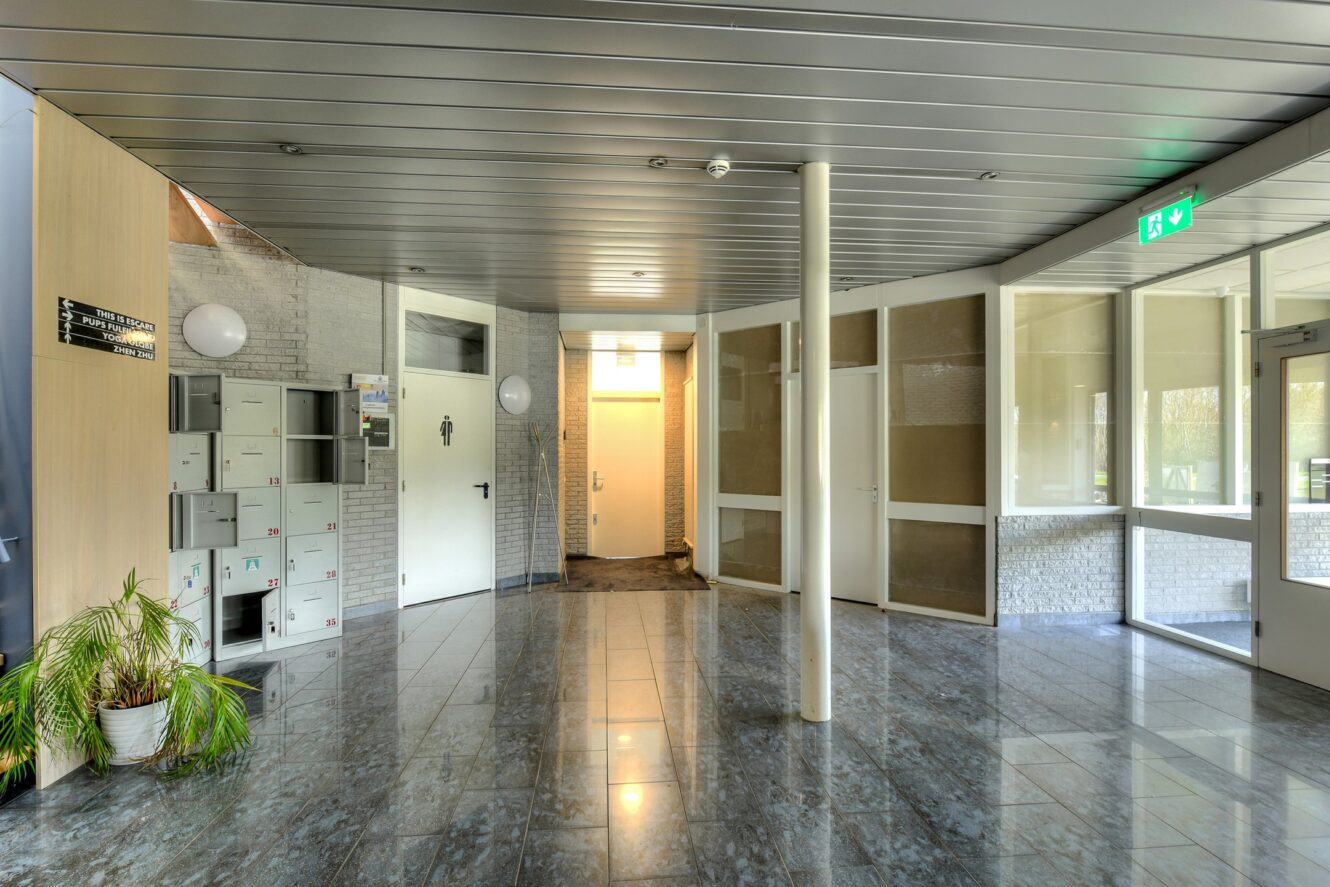 Een schone, moderne hal met betegelde vloeren, kluisjes, een plant in potten, glazen muren en een bord met een nooduitgang boven een deuropening.