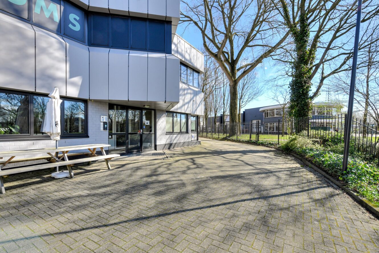 Modern gebouw aan de buitenkant met grote ramen, een witte picknicktafel en een geplaveide binnenplaats; bomen en een ander gebouw zichtbaar op de achtergrond op een heldere, zonnige dag.