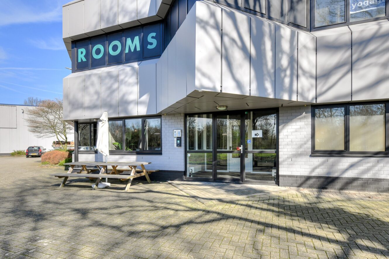 Buitenaanzicht van een modern gebouw met een bord ROOMS, glazen toegangsdeuren, een picknicktafel en een parkeerplaats op de achtergrond.