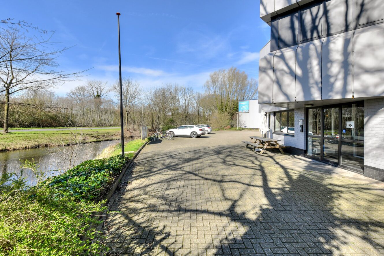 Een verharde parkeerplaats naast een kanaal met twee geparkeerde auto's, een fiets en een ingang van een gebouw onder een zonnige hemel met bomen op de achtergrond.
