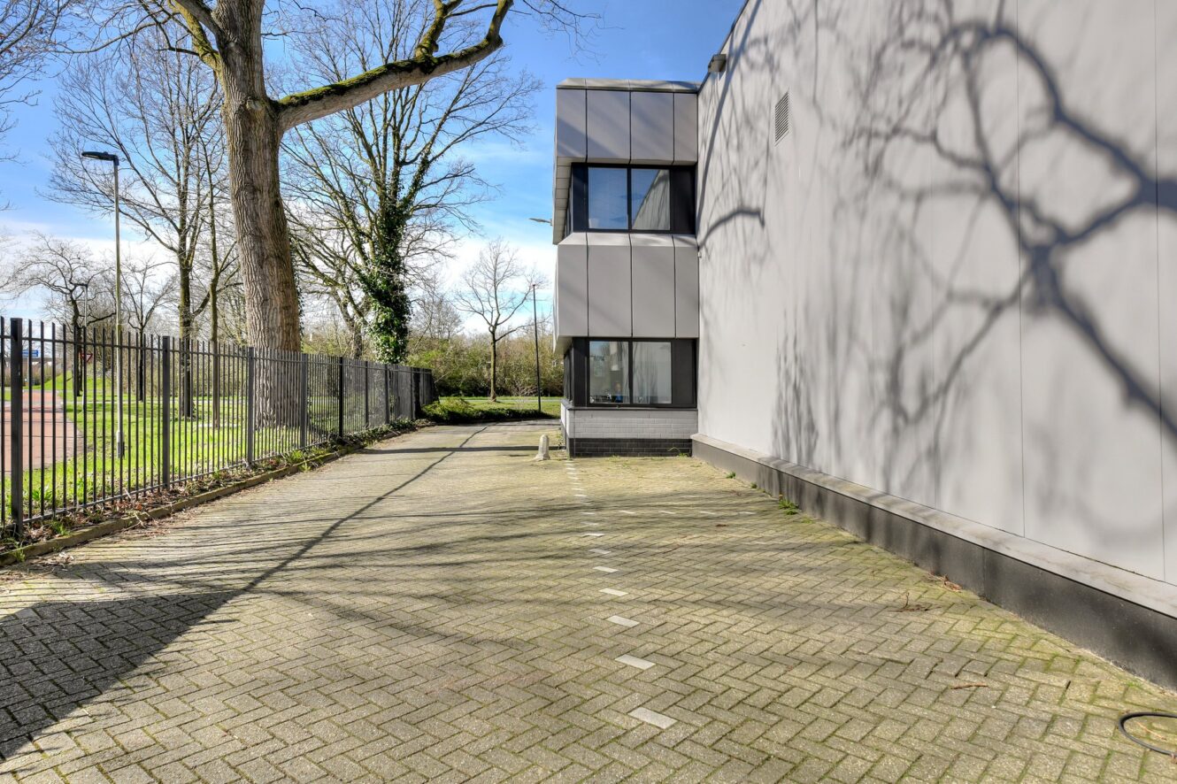 Een geplaveid pad loopt langs een modern gebouw met grote ramen, omzoomd door een metalen hek en bladerloze bomen die schaduwen op de grond werpen.