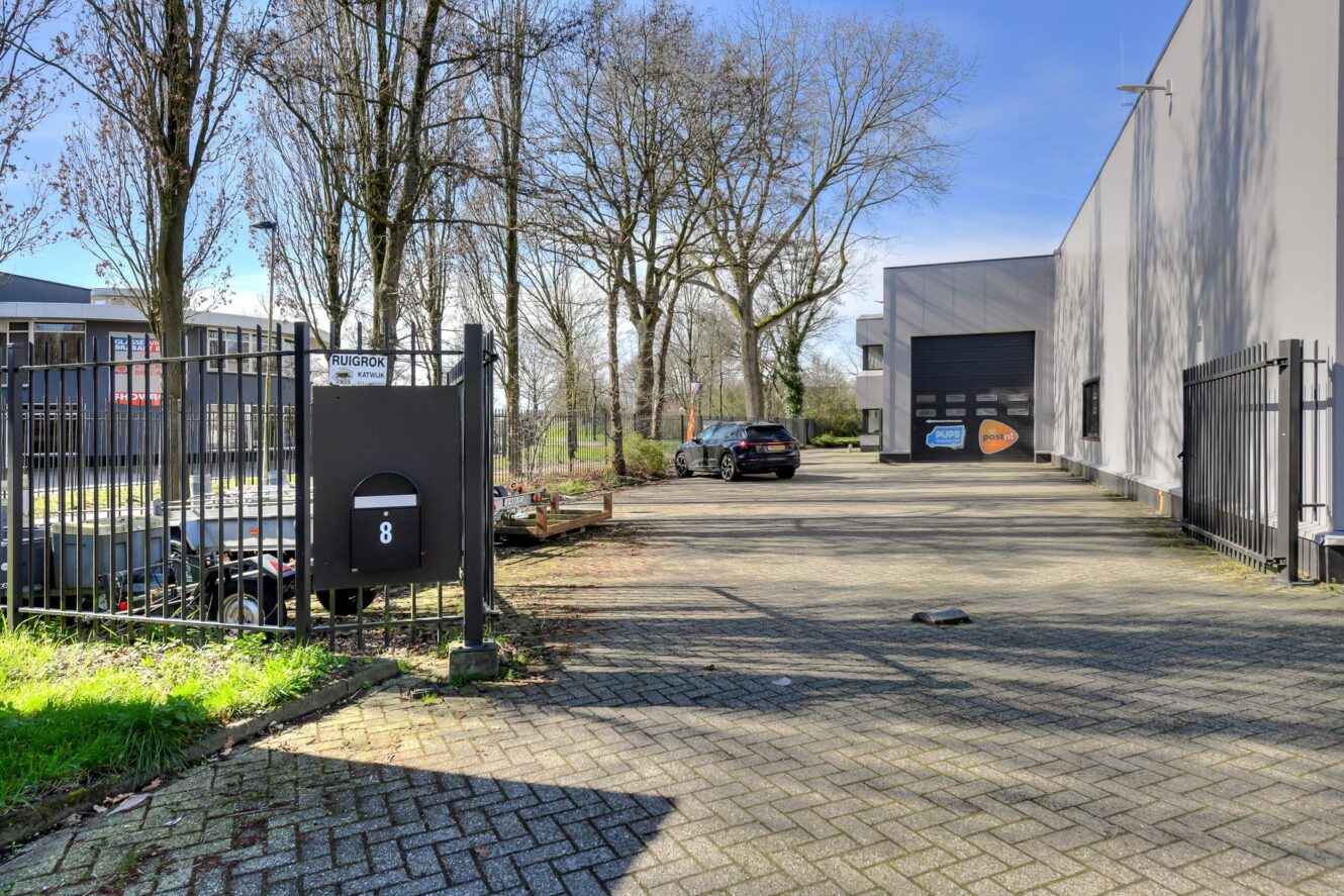 Afgesloten ingang van een commercieel pand met een brievenbus met het opschrift 8, geparkeerde auto's en een magazijngebouw op de achtergrond. Bomen omzomen het gebied onder een heldere hemel.