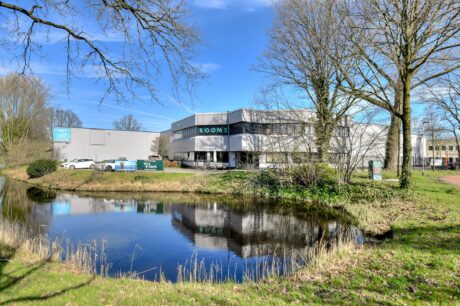 Modern kantoorgebouw met grote ramen en een ROOMS-bord, omringd door bomen en gras, weerspiegeld in een kleine vijver onder een helderblauwe hemel.