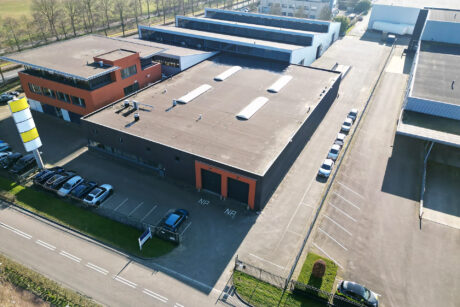 Luchtfoto van een industrieel gebouw met een plat dak, aangrenzend parkeerterrein met auto's en omliggende verharde gebieden en wegen.