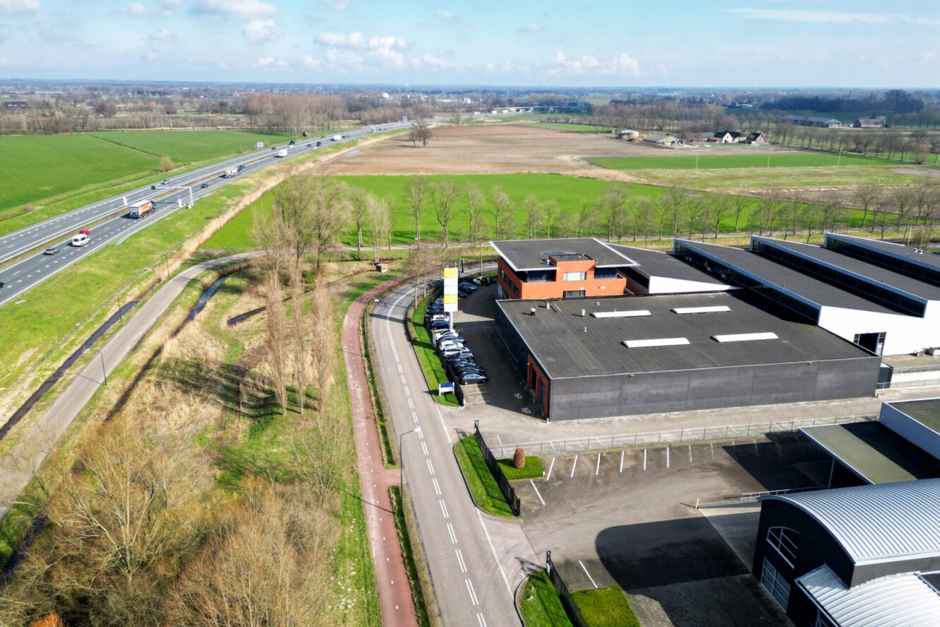 Luchtfoto van een industrieel gebouw naast een weg en fietspad, met buiten geparkeerde auto's en velden en een snelweg op de achtergrond.