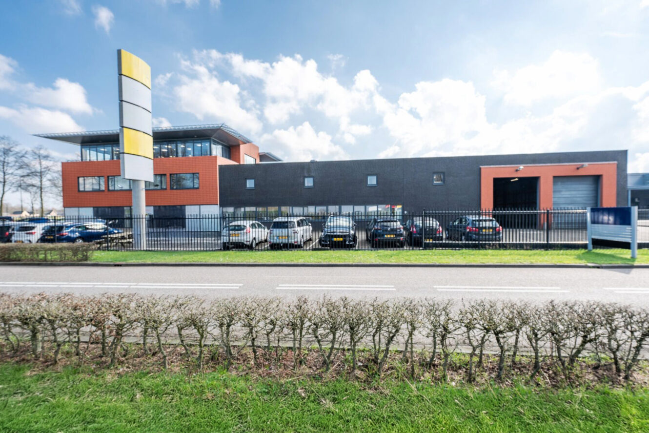 Modern industrieel gebouw met oranje en grijze gevel, omheind parkeerterrein met auto's en een groot bord bij een weg op een gedeeltelijk bewolkte dag.