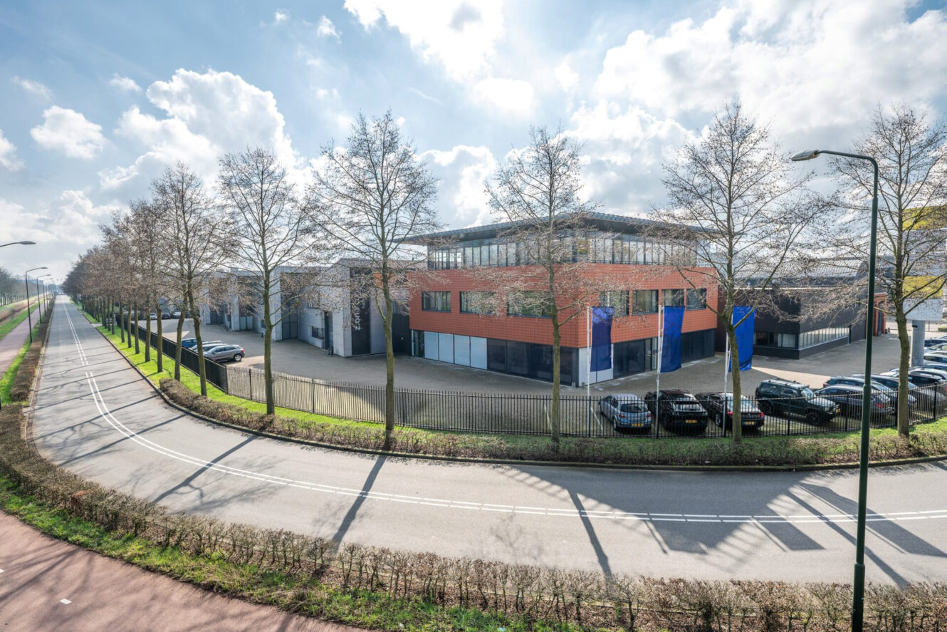 Een modern industrieel gebouw met oranje en blauwe panelen, omringd door bladerloze bomen, parkeerplaatsen en een bochtige weg onder een gedeeltelijk bewolkte hemel.