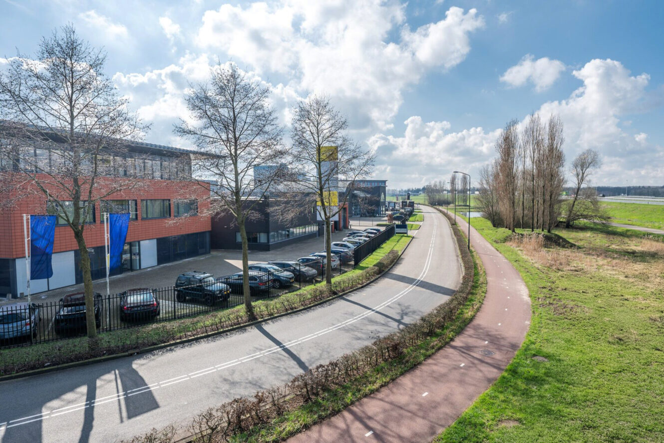 Een tweebaansweg met een parallel fietspad loopt naast een kantoorgebouw met geparkeerde auto's; open velden en bomen omzomen de overkant onder een gedeeltelijk bewolkte hemel.