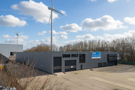 Een groot industrieel gebouw met meerdere garagedeuren, omringd door bomen, met windturbines en een gedeeltelijk bewolkte lucht op de achtergrond.