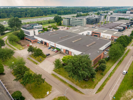 Luchtfoto van een industrieel complex met grote pakhuisachtige gebouwen, parkeerplaatsen en omliggend groen naast een snelweg.