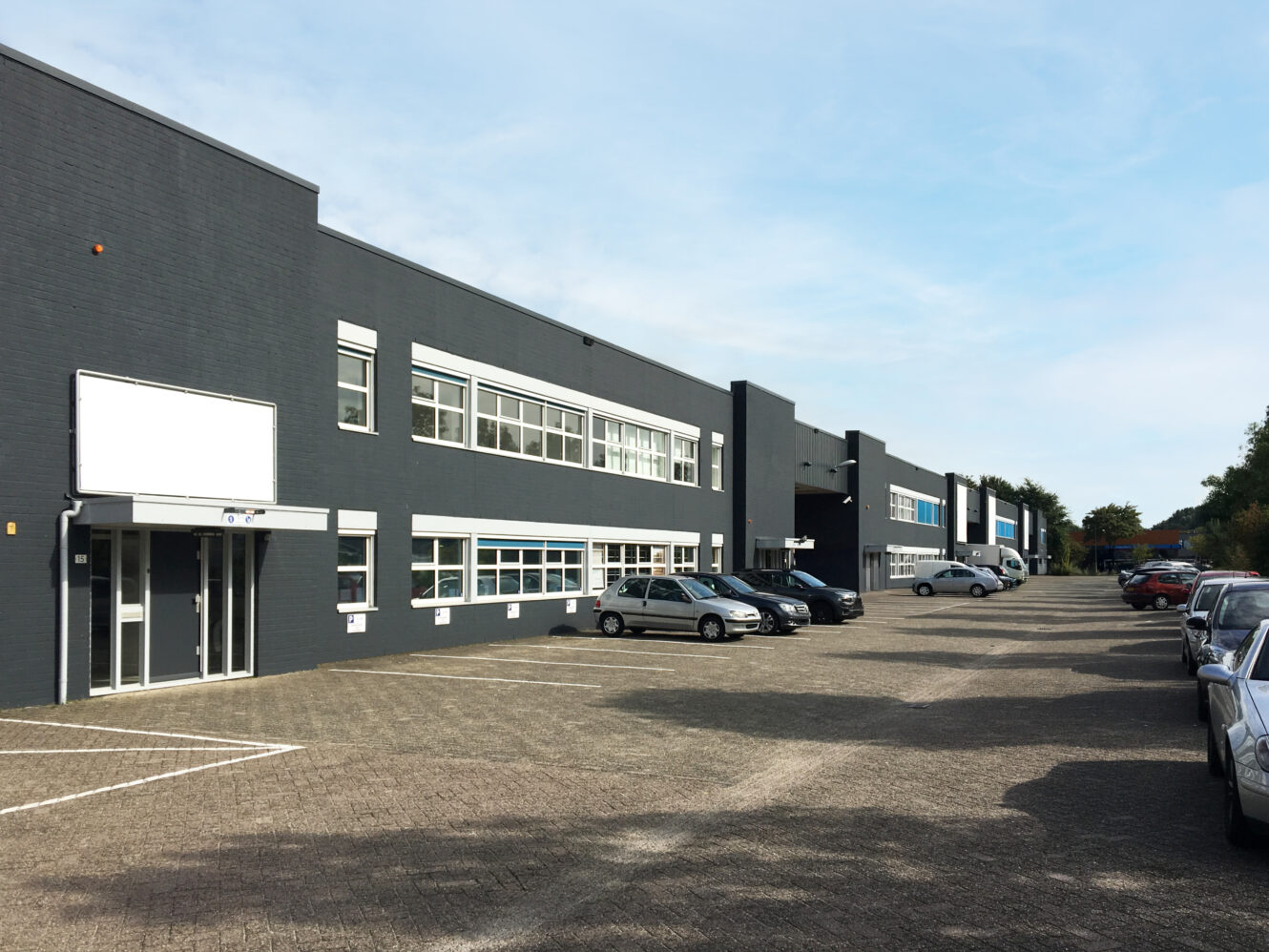 Een rij moderne industriële gebouwen met grote ramen en geparkeerde auto's op een ruim geplaveid terrein onder een heldere hemel.
