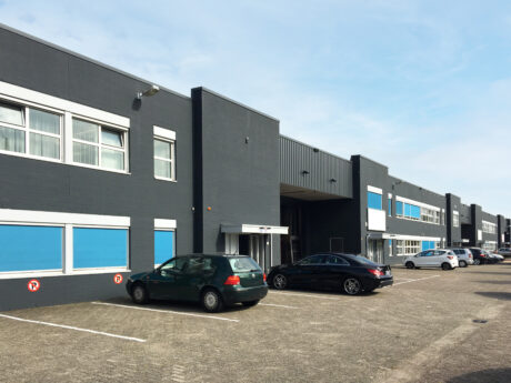 Modern industrieel gebouw met donkergrijze buitenkant, meerdere ramen en blauwe accenten. Verschillende auto's staan geparkeerd langs een verharde parkeerplaats onder een heldere hemel.